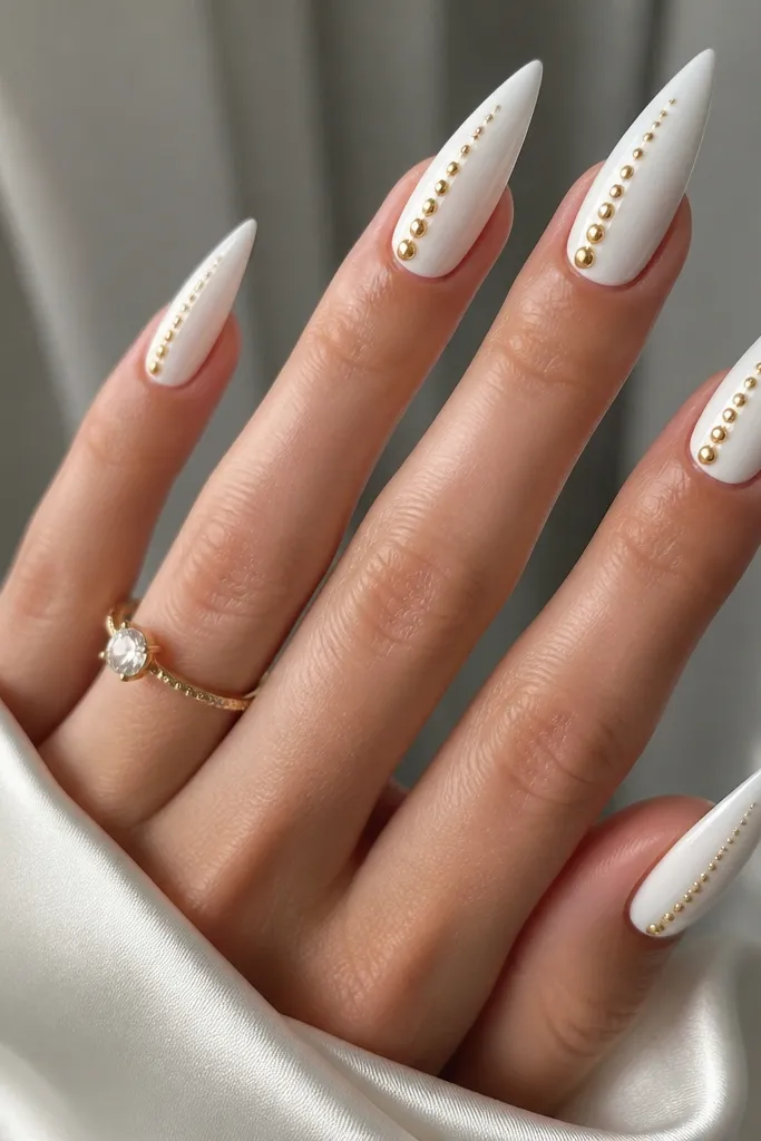 Glossy white stiletto nails with a single row of tiny gold studs along one side near the middle. The studs are evenly spaced and taper slightly as they approach the tip.