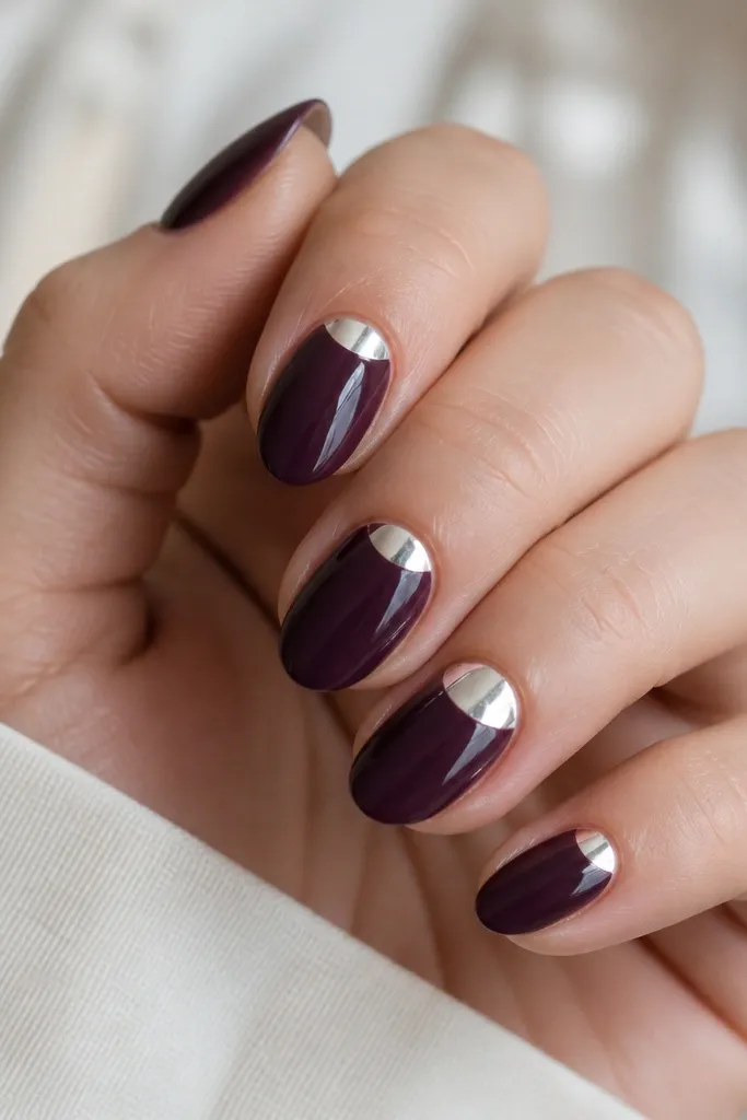 Short almond nails painted a deep grape jelly purple with a glossy finish. On each nail, a clean silver foil half-moon sits at the cuticle, leaving the rest of the nail solid purple. One accent nail has slightly larger foil at the half-moon.
