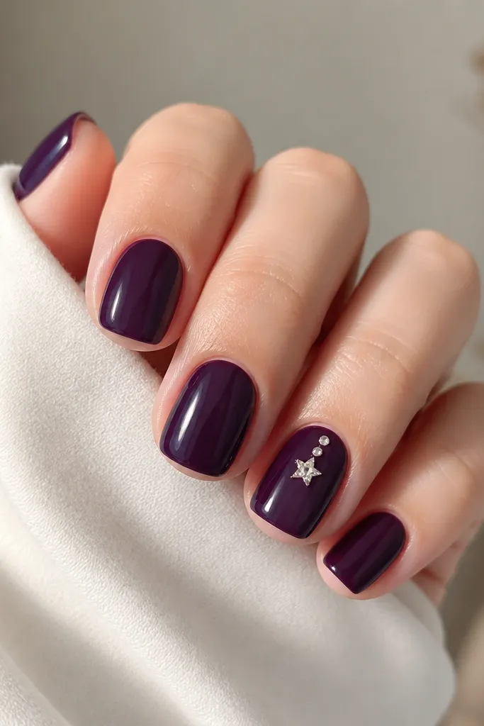 Short squoval nails in a glossy dark purple. Four nails are solid color; one accent nail has a small star-shaped rhinestone cluster near the cuticle, with two tiny clear stones and one larger star stone.