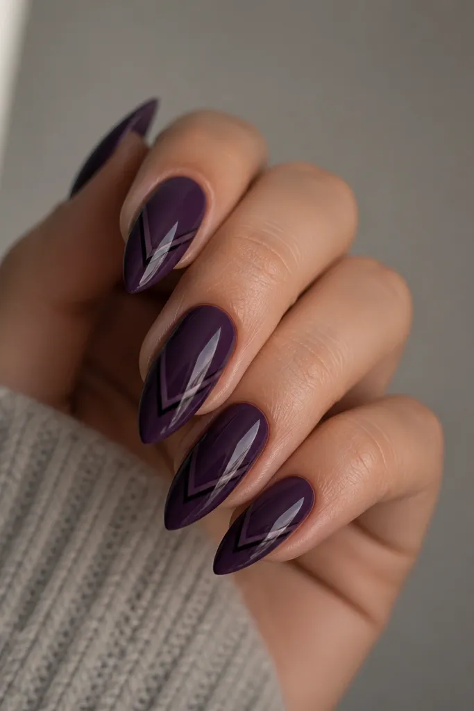 Long almond nails with a glossy dark purple base. On each nail, a black V-shaped outline sits in the center, pointing toward the tip, with thin lines and sharp edges. The rest of the nail stays solid purple.