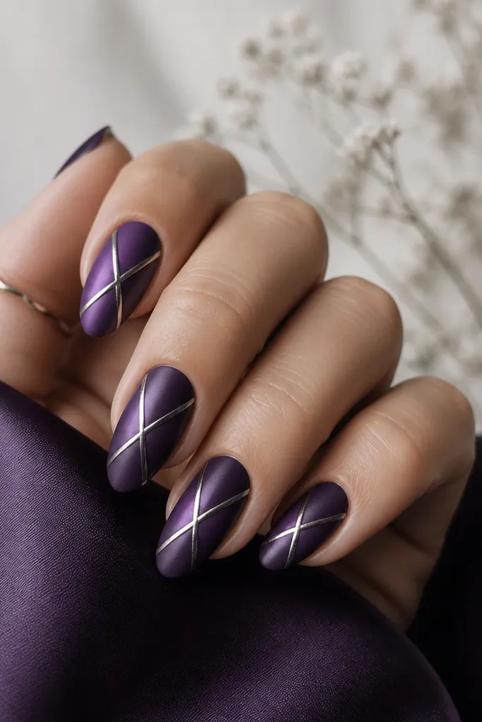Medium almond nails in a satin, not fully matte, dark purple finish. Silver lines crisscross near the center of each nail forming a loose X pattern. The nails look soft with a slight sheen.