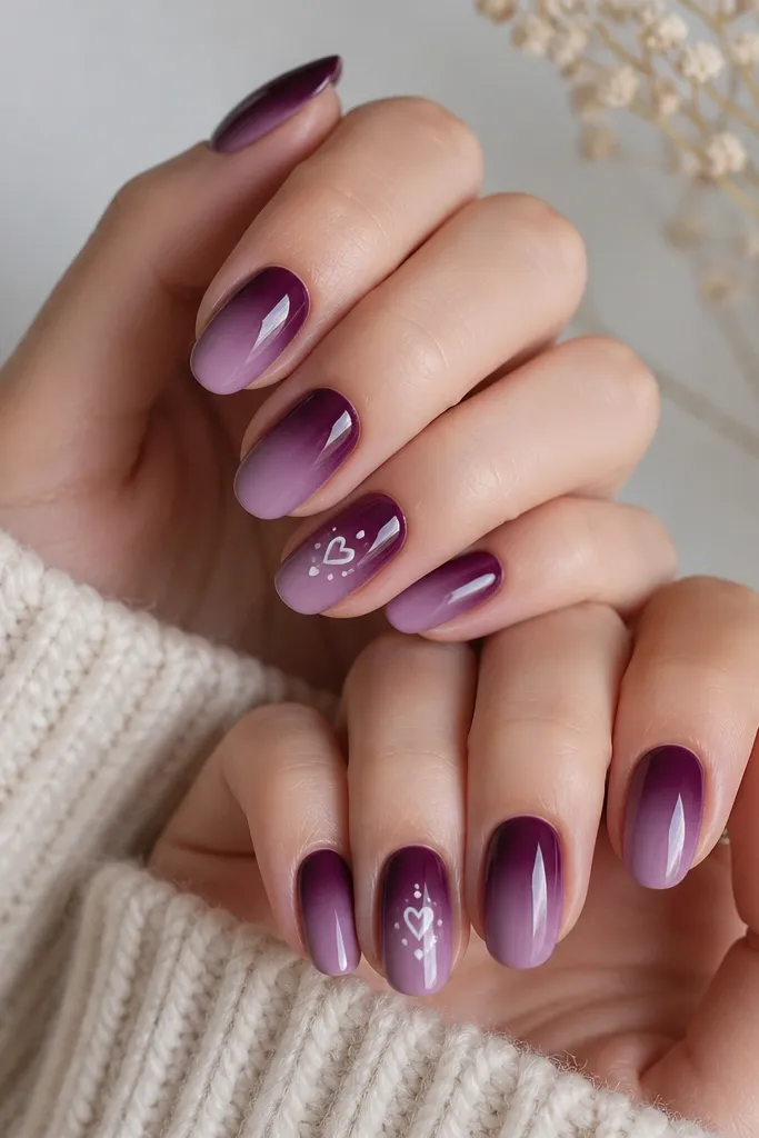 Medium oval nails with a plum ombré that fades from deep plum at the cuticle to a lighter lilac toward the tip. Two accent nails have tiny white hearts placed near the center, with one heart per nail and a couple small dots around it. Glossy finish.