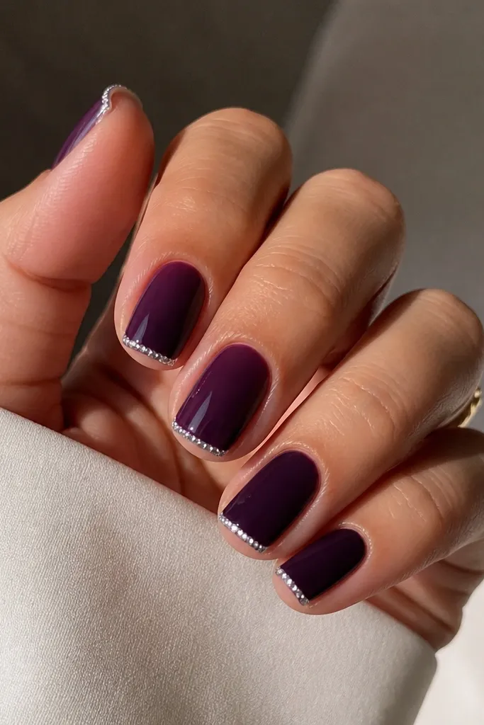 Short squoval nails in glossy dark purple. Each nail has a classic French tip, but the tip is a thin white line. Along the edge of the white tip, a line of tiny clear rhinestones forms a micro border. The rest is solid purple.
