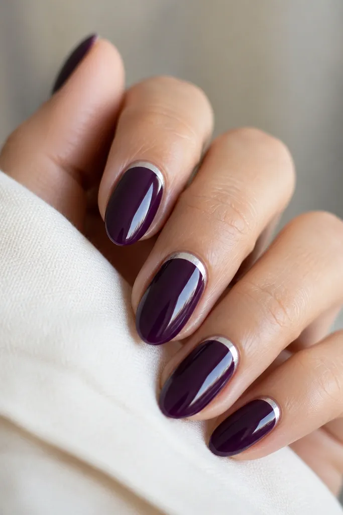 Short almond nails painted deep eggplant purple with a glossy topcoat; a thin silver foil crescent sits right at the cuticle line on each nail, leaving the rest solid purple.
