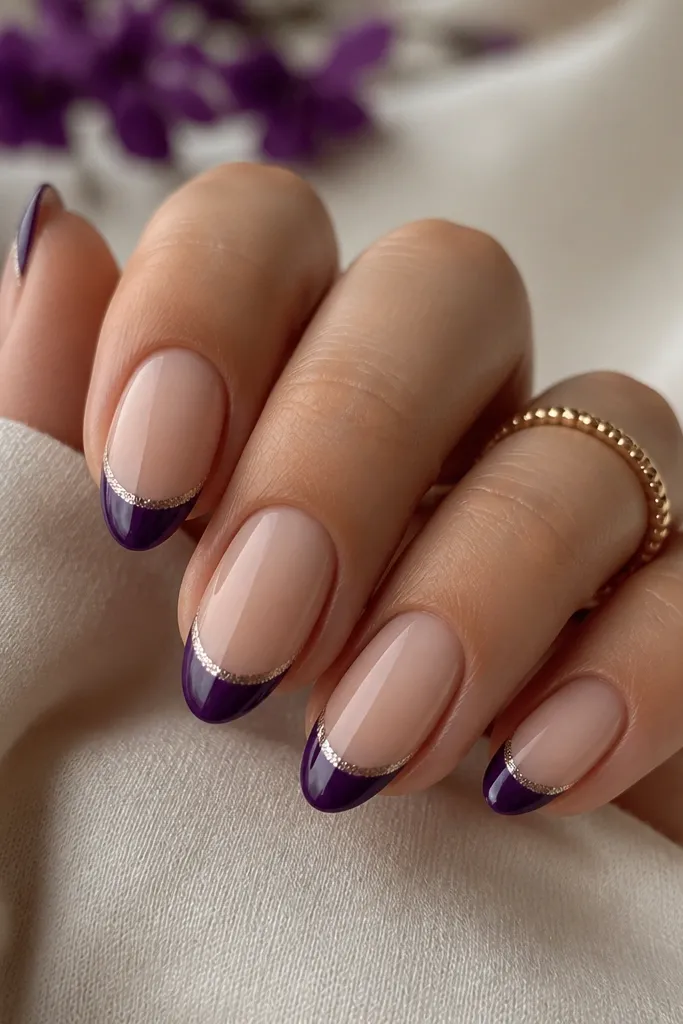 Short almond nails with a natural nude base; crisp dark purple French tips and a thin micro-glitter band between the nude and purple tip.