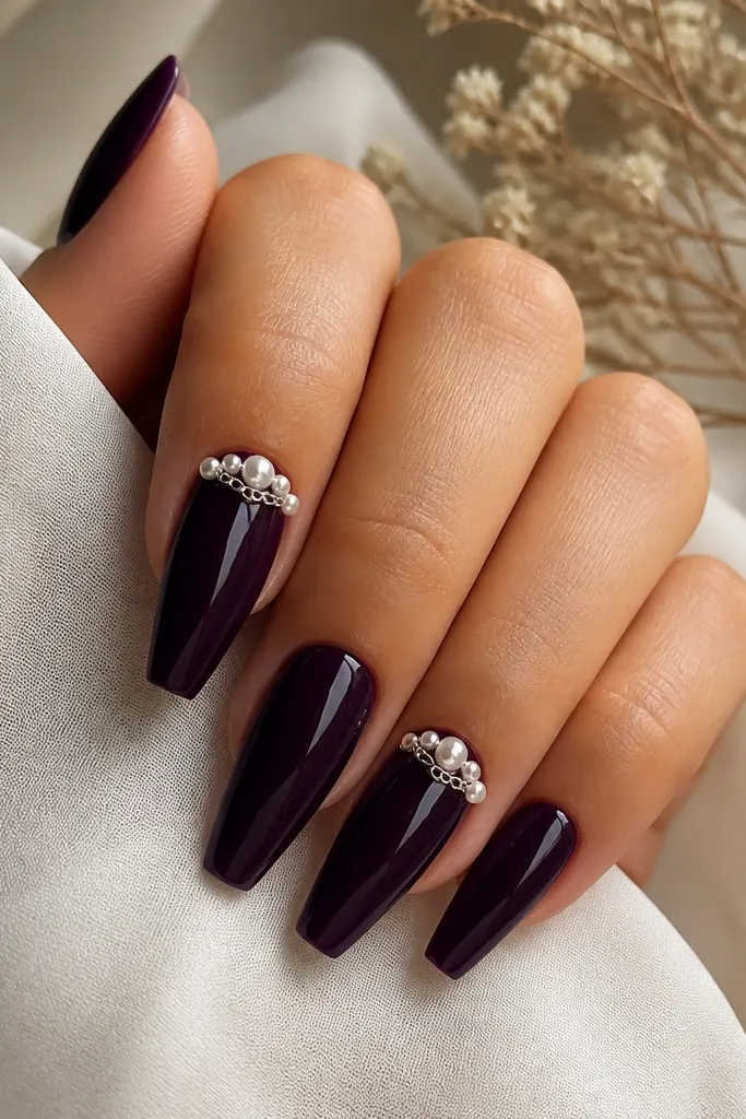 Coffin nails painted deep dark purple glossy; two nails have small pearl clusters and tiny silver chain links arranged diagonally near the cuticle, other nails plain.