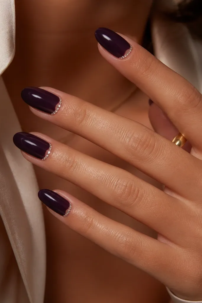 Medium almond nails in glossy dark purple; small clear rhinestones form a thin frame around the cuticle area, with a clean gap in the center.