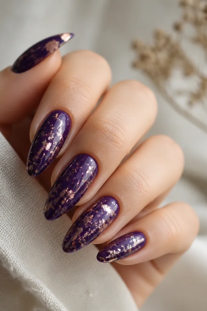 Almond nails painted glossy dark purple; scattered rose-gold foil flecks sit randomly across the surface, especially near the center and tips.