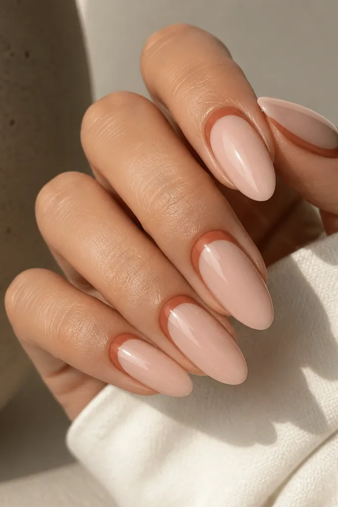 Almond nails with a nude center panel. Terracotta reverse French arcs at the cuticle, leaving the middle nude. Glossy finish.