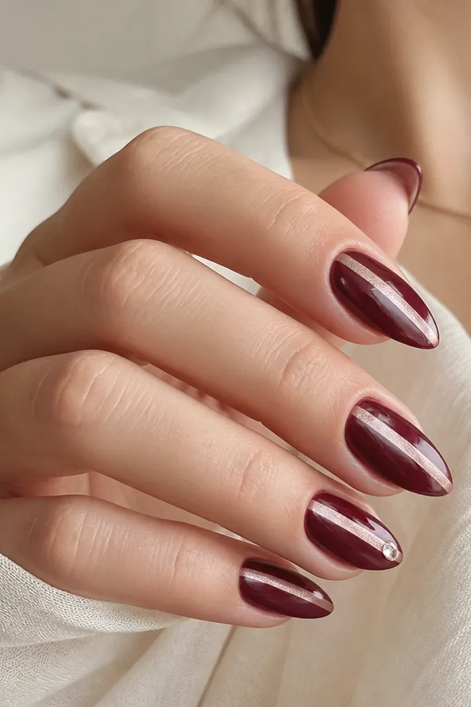 Medium almond nails in glossy burgundy wine. Each nail has a narrow pearlized stripe down the center in pale champagne. One nail has a tiny crystal at the tip.