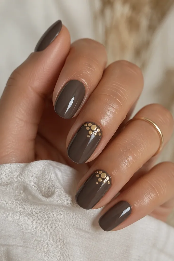 Charcoal gray-brown nails with a glossy finish. Two nails have warm gold dots clustered near the cuticle like tiny coins. Other nails are solid color.