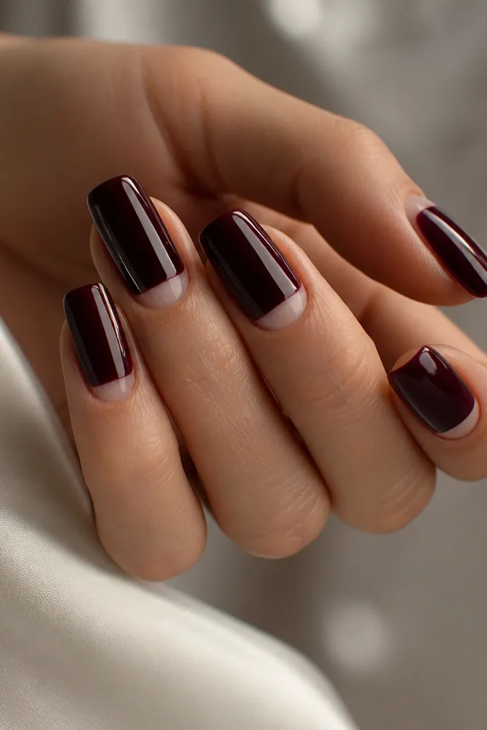 Long squoval nails painted black cherry glossy. At the cuticle, a small crescent of natural nail is left bare, creating negative space. One nail has a thin silver line along the side.