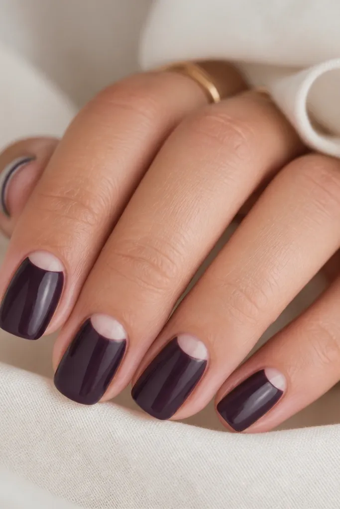 Short squoval nails painted smoky eggplant purple. Each nail has a crisp negative-space half-moon near the cuticle in the natural nail color, separated by a clean curved line.