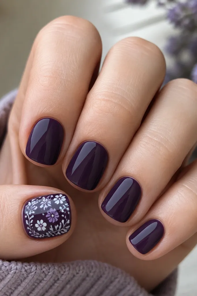 Short nails painted solid dark purple. The thumb nail has a small floral stamp pattern in light purple and white, with crisp stamped lines and a glossy finish.