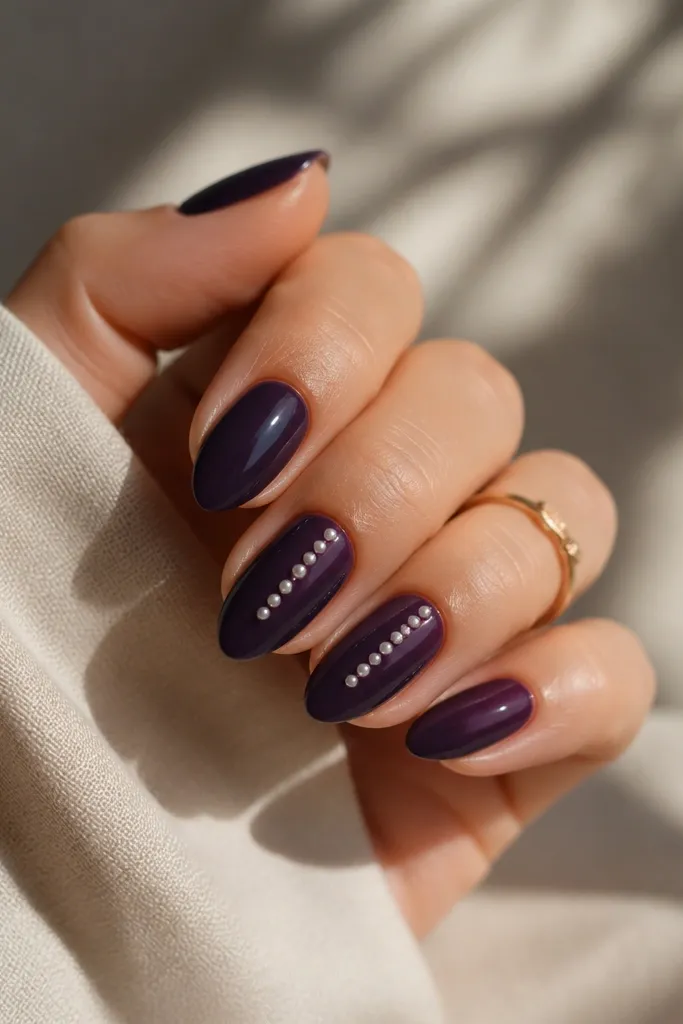 Short almond nails in dark purple gloss. Small pearl-like dots sit in a vertical row near the center on two nails, catching light like tiny beads.