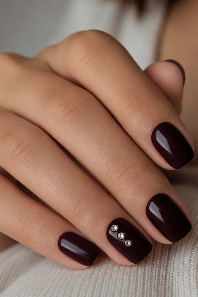 Deep near-black plum nails with three tiny crystal dots on the ring finger. Crystals are faceted and catch light; other nails are solid.