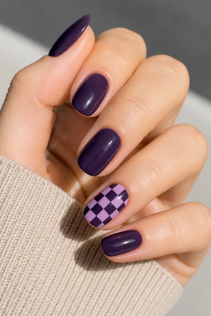Solid dark purple nails. One accent nail has a small checkered pattern in lighter lavender and dark purple squares, arranged evenly.