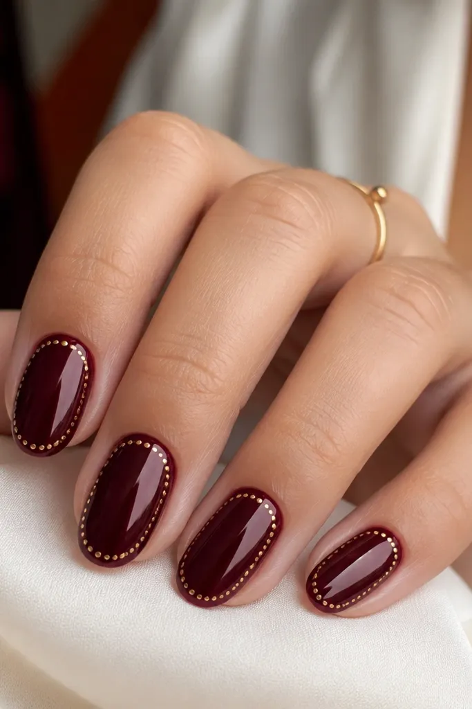 Burgundy glossy nails; a thin border of tiny gold dots runs along the nail edge near the tip, like a dotted frame; one accent nail has the border only on one side.