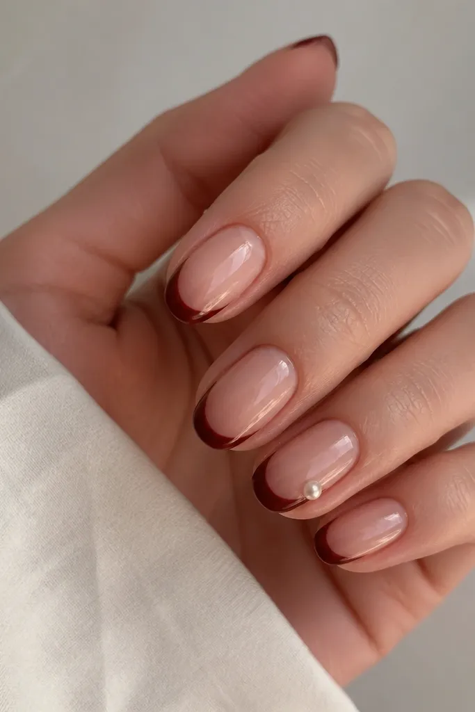 Nails have a nude base. French tips are rosewood brown-red. On one nail, a single tiny pearl sits on the side near the tip curve, glossy overall.