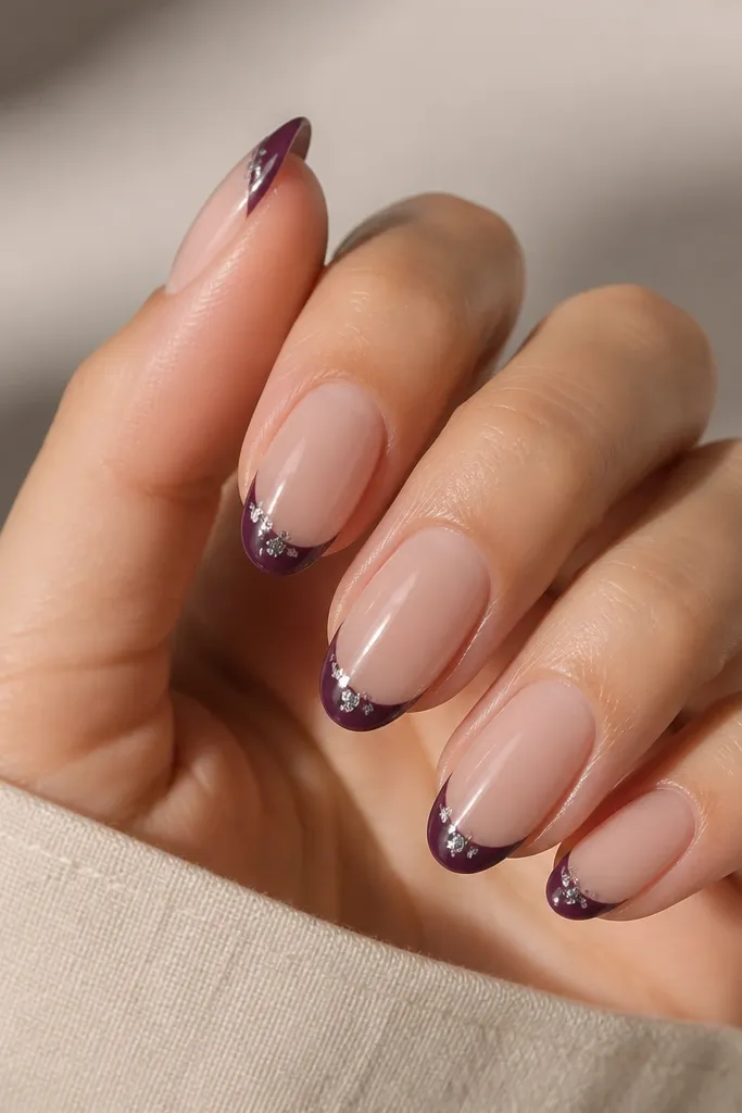 Nails have a nude base and glossy finish. The French tips are plum purple. On each tip's outer corner, there are small silver foil flakes pressed into a clear gel spot.