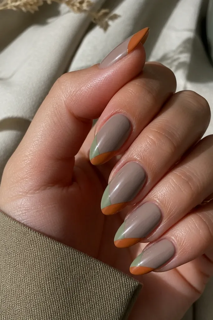 Nails have a moody gray-nude base. The French tips are split diagonally into sage green and rust orange, with a sharp diagonal line. Glossy top coat makes the colors crisp.