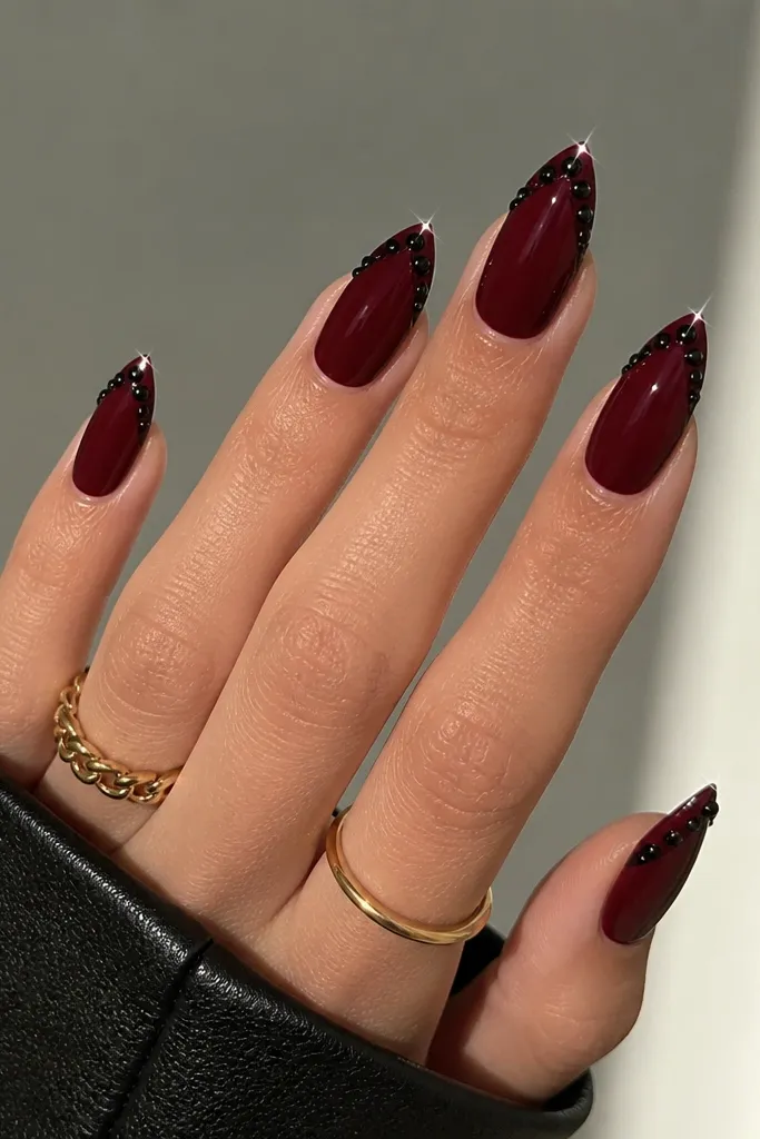 Oxblood nails with a V-shaped French tip. The V is outlined in black, and tiny black rhinestones sit on the V line like spaced studs. Glossy finish with sparkle at the tips.