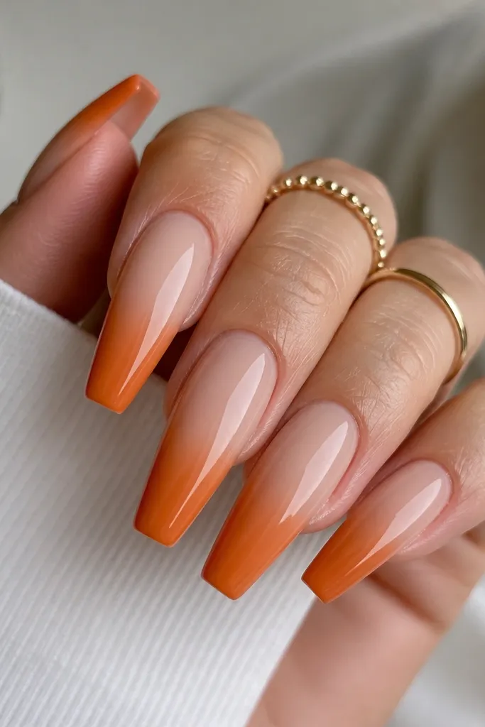 Long coffin nails with a smooth ombre. The base is warm nude, and the tips fade into opaque pumpkin orange. The gradient is soft, with no harsh line. Glossy finish reflects light.