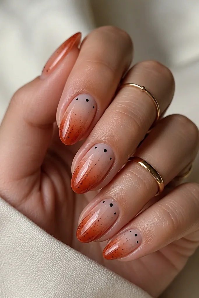 Medium oval nails with a burnt orange gradient at the tips fading into nude. Over the gradient are tiny charcoal dots scattered like stars, with a few slightly larger dots near the cuticle area. Glossy finish.