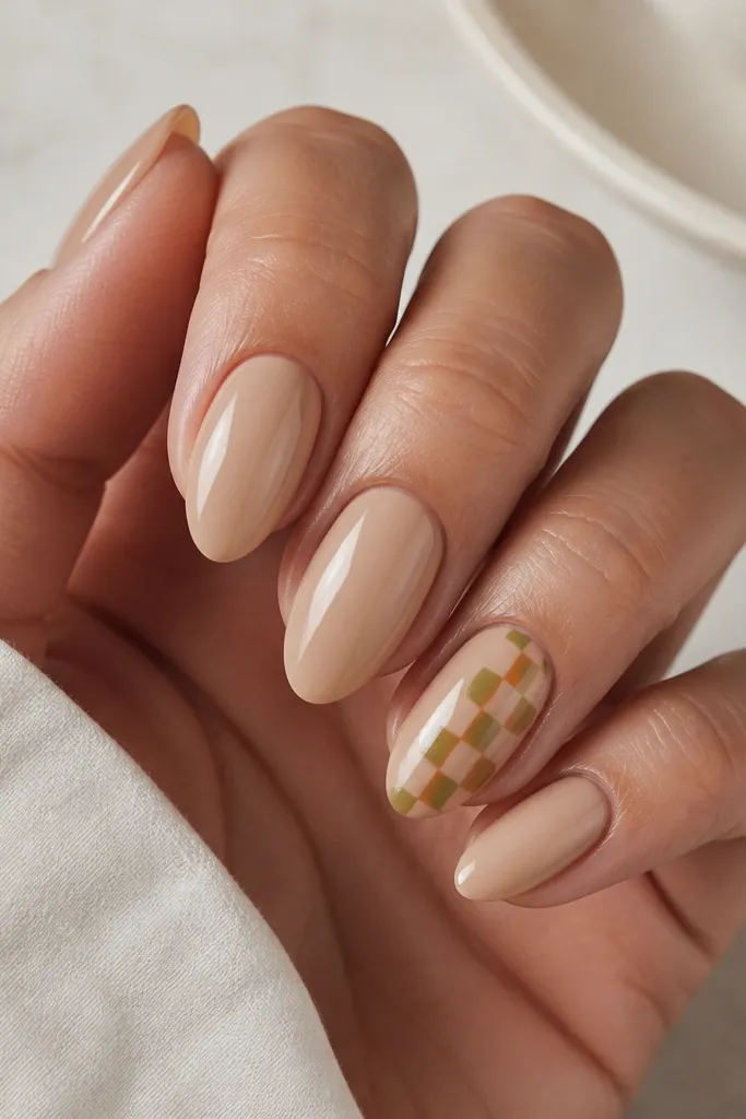Medium almond nails with solid warm nude base. On the ring finger nail, there is a diagonal checker pattern made from alternating orange and olive-green squares. The edges are crisp, and the nail is glossy.