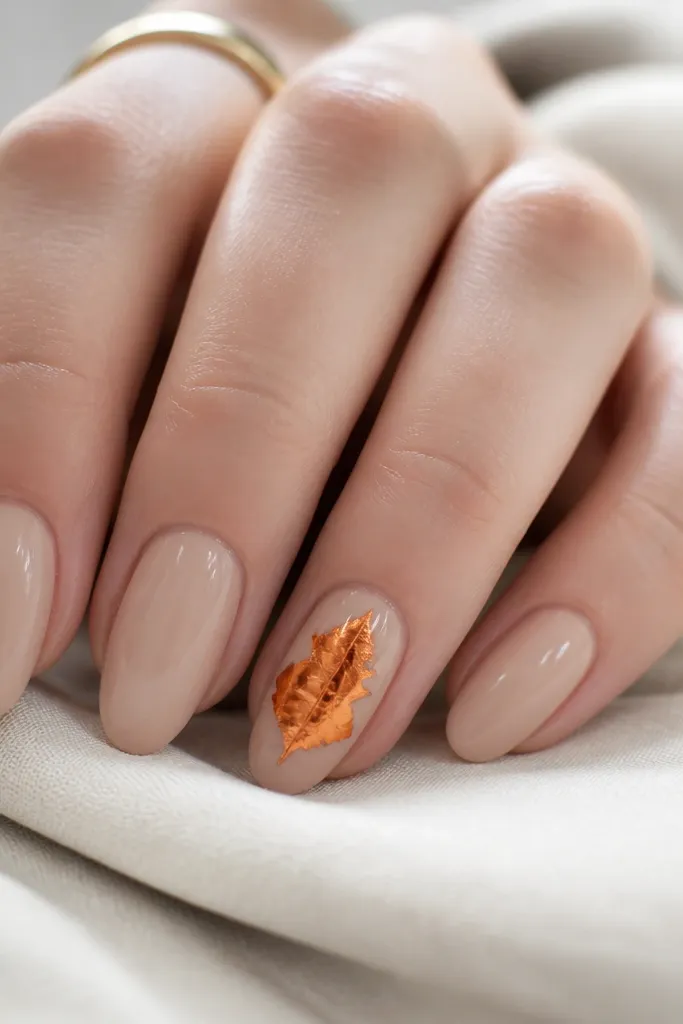 Medium oval nails with solid nude base and glossy finish. One accent nail has a pumpkin orange foil leaf shape pressed across the center. The foil has a metallic shine with slight wrinkles. Other nails stay plain.