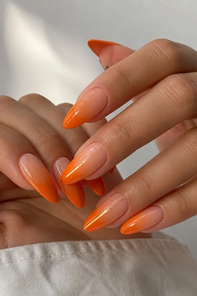Long almond nails with a tangerine gradient from nude at the base to bright orange at the tips. Over the gradient, there are two thin gold micro lines running vertically on two nails. The rest are solid gradient. Glossy top coat.
