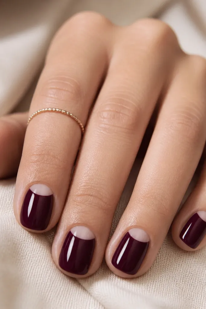 Short nails with deep plum color covering most of the nail. At the cuticle, there is a reverse half-moon window of clear nude, separated by a crisp curved edge. Glossy finish throughout.