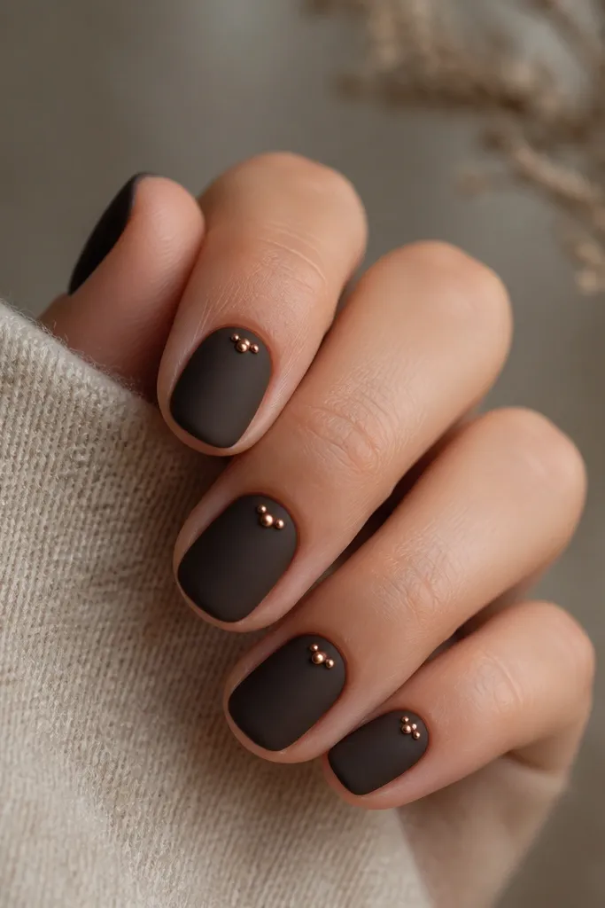 Short nails in matte blackened brown. On each nail, there is a small cluster of copper dots near the cuticle, like three tiny beads. The dots are glossy copper against the matte background.