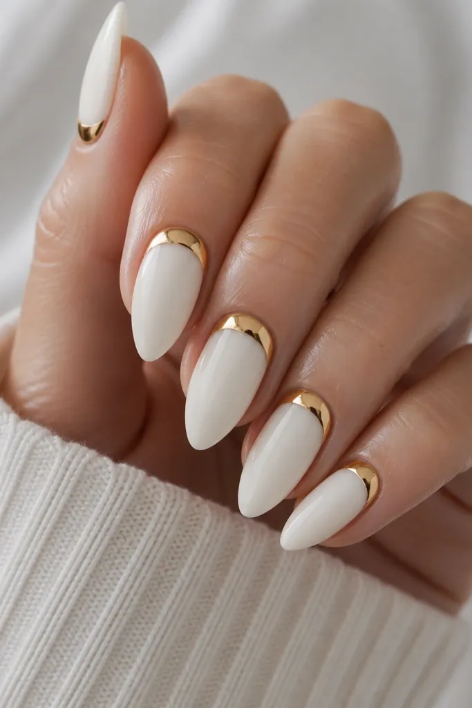 Long almond nails painted milky porcelain white with a glossy mirror gold half-moon at the base of each nail, centered and clean, with a high-shine top coat.