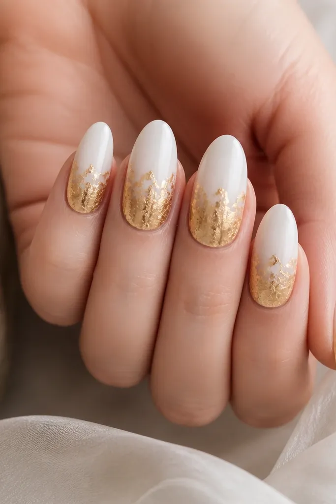 Milky white almond nails with gold leaf covering the lower half from mid-nail to tip, leaving the top half clean white, glossy top coat over leaf.