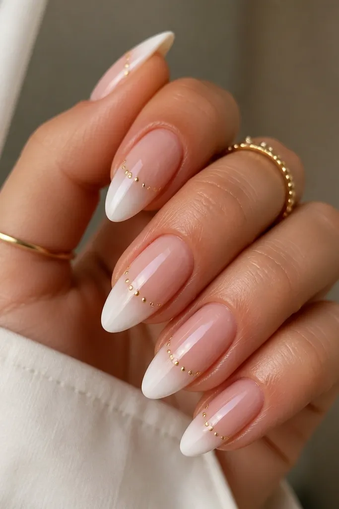 Almond nails with a white ombré that gets stronger at the tips, and small gold studs spaced in a line across the tip area, glossy top coat.