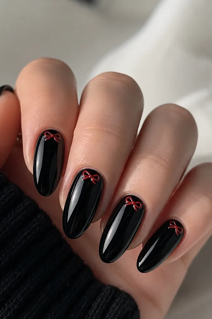 Nails are glossy black. Near the cuticle, there's a small red bow tie shape with two loops and a knot, plus a tiny white highlight at the top of each loop.