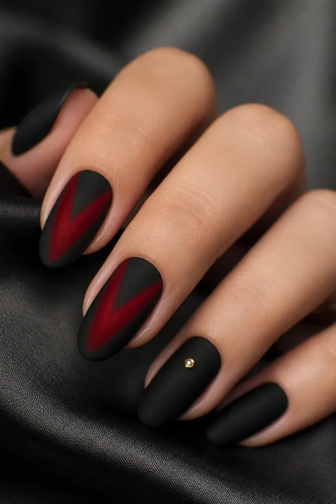 Most nails are matte black. On the index and middle nails there is a V-shaped red panel that looks velvety, with a soft gradient at the edges. The ring finger has a single small gold stud at the center of the matte black nail.