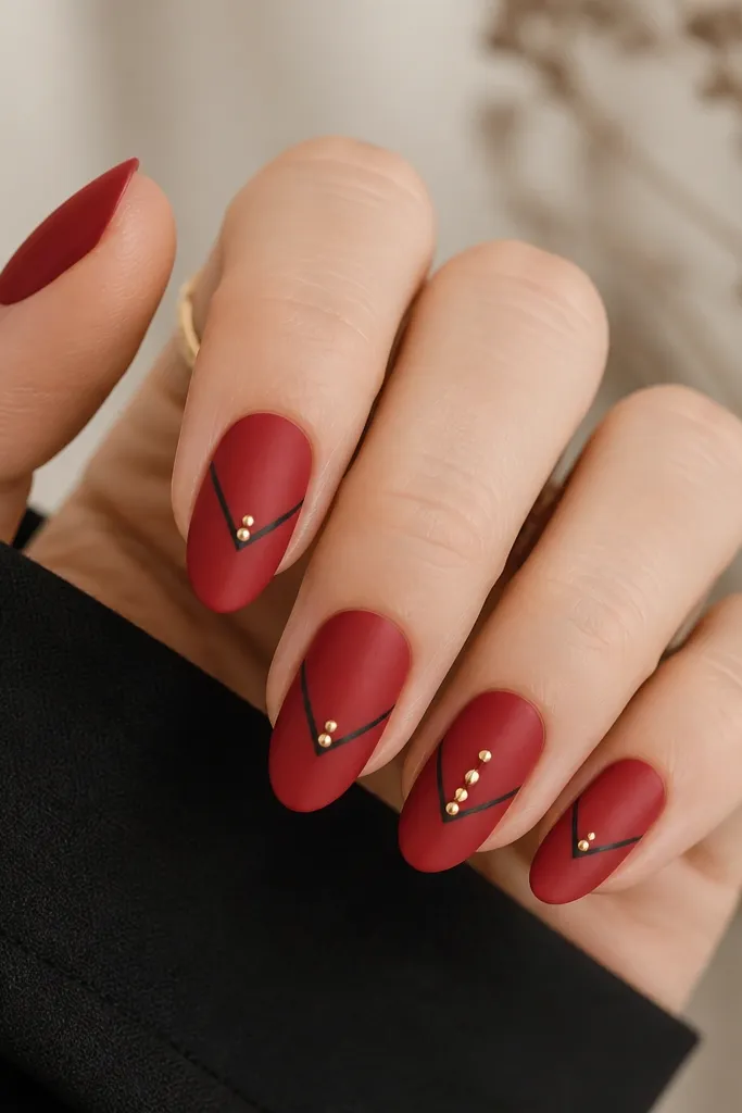 Nails have a satin-like red finish, not fully mirror. A black chevron points toward the center of each nail. Each nail has two tiny gold micro studs placed along the chevron arms, with the ring finger having three studs.