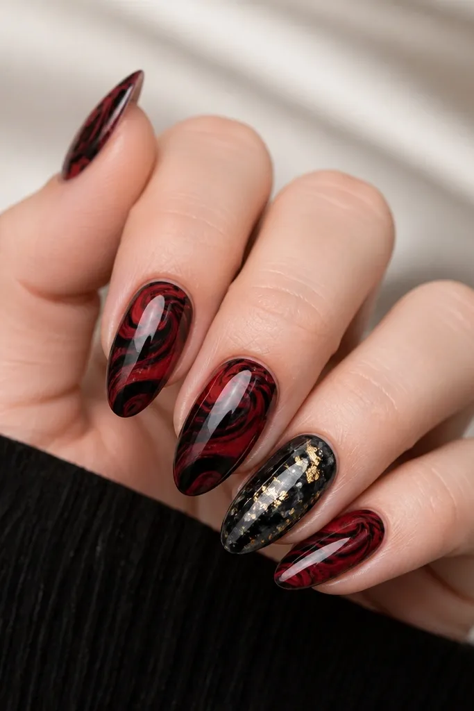 Most nails show red and black marble swirls swirling diagonally. The ring finger is mostly black marble with scattered gold leaf shards across the center. All nails have a glossy finish.