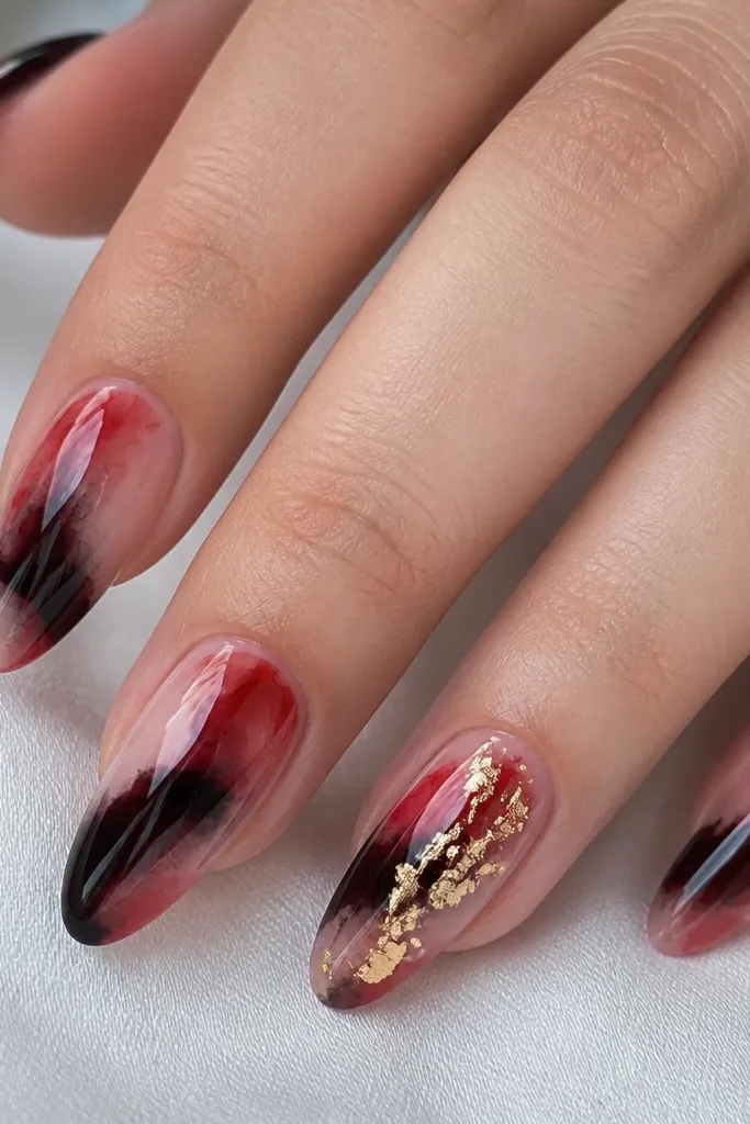 Nails show watercolor-like swipes of red and black that blend softly, not solid blocks. The ring finger has gold leaf pieces drizzled diagonally across the center. All nails are glossy.