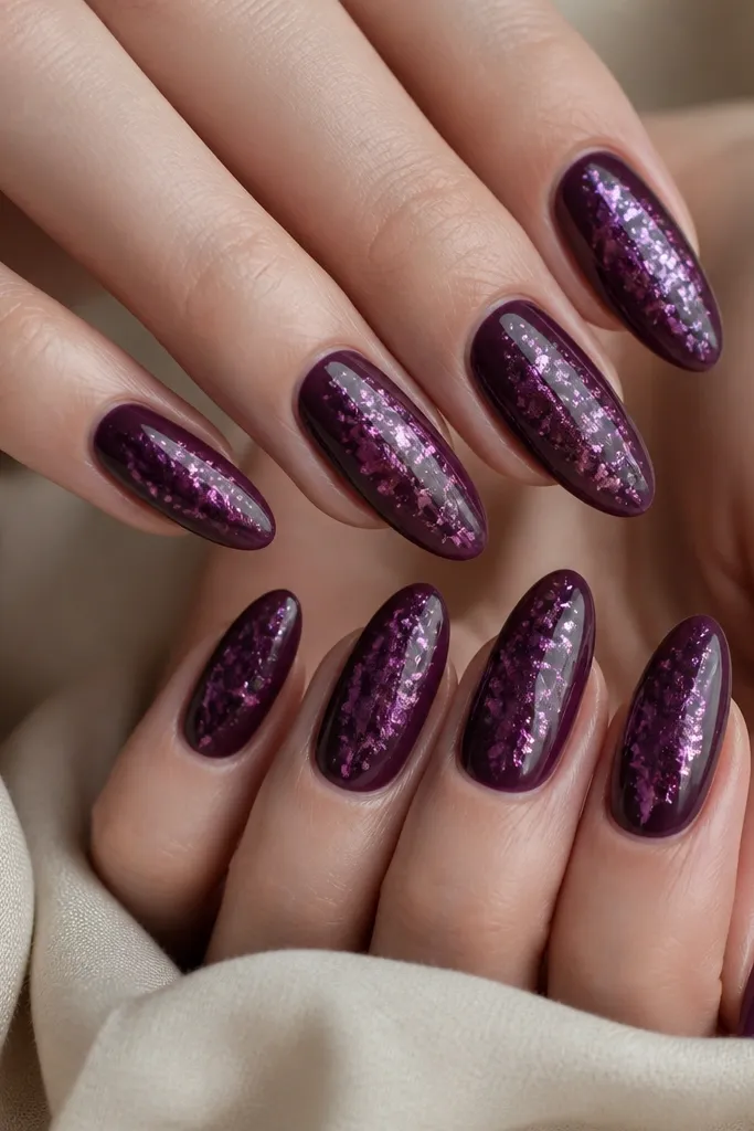 Almond nails with deep plum base; irregular plum foil flakes are placed near the center; a sheer layer of fine glitter veil sits over the flakes, glossy finish.