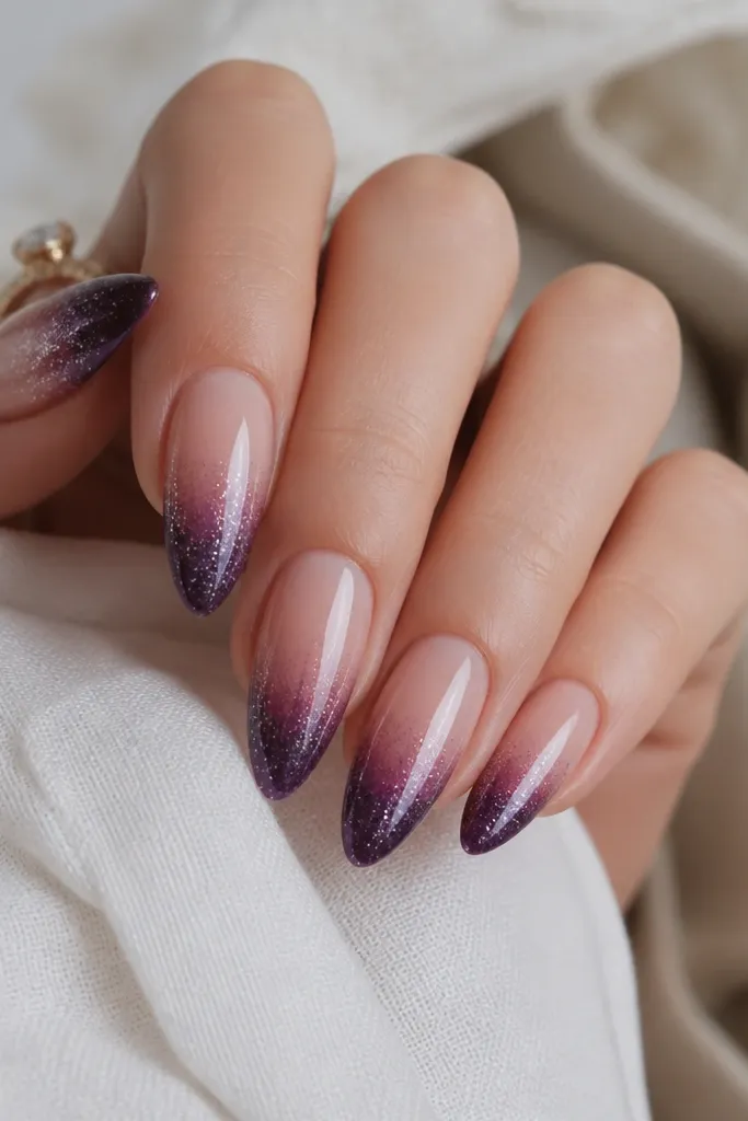 Long almond nails with dark purple tips fading into a sheer mid-nail; glitter is concentrated at the extreme tips in a smooth gradient, glossy finish.
