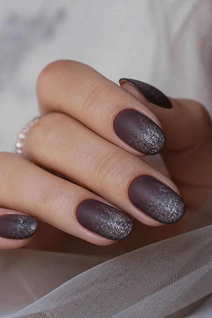 Short almond nails painted a smoky eggplant base with a soft, airbrushed haze. Silver glitter fades from the center of each nail toward the tips, leaving the cuticle area matte and velvety. Subtle gray smoke wisps blur the edges so it looks like pigment drifting under glass.