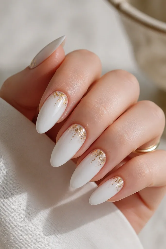 Long almond nails painted in a thick, glassy white gel. Near the cuticle, small gold flakes are scattered like a gentle sprinkle, with most flakes clustered around the center.