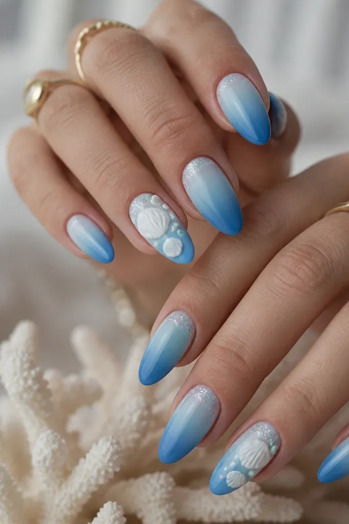 Long almond nails with a blue ombre from sky at the cuticle to deeper ocean at the tip. The cuticle area has a shimmering gel melt. Accent nails include raised shells in white with tiny seafoam dots.