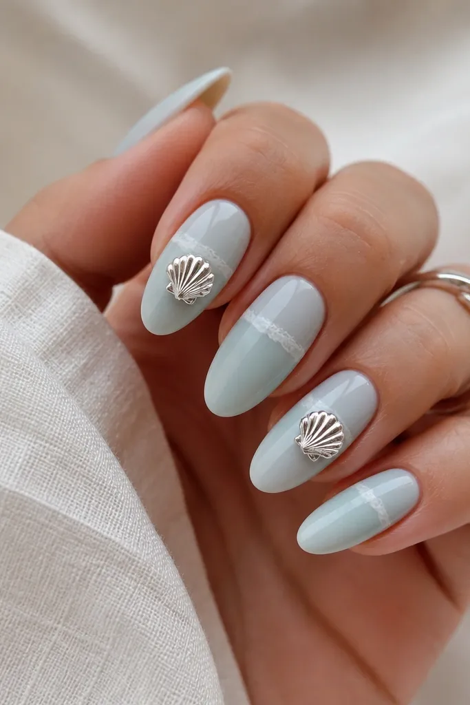 Almond nails painted frosted icy blue. Accent nails have silver chrome seashell charms with a mirror-like ridge texture. A thin frosted waterline runs across the middle on each nail.