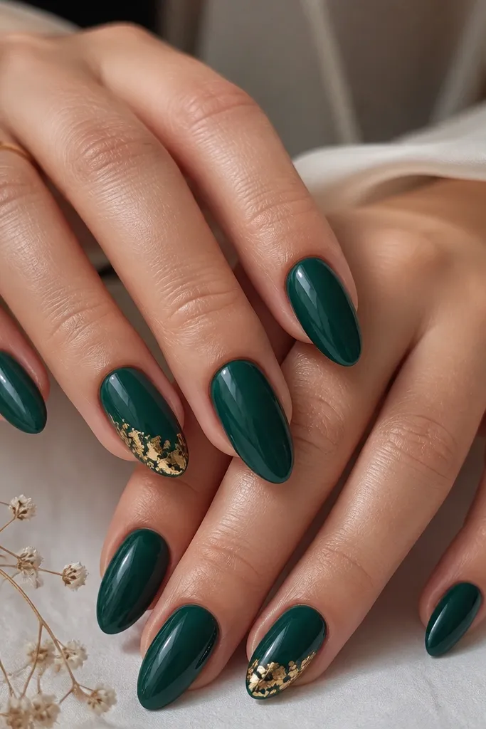 Almond nails in glossy emerald green with a few nails featuring small irregular gold leaf pieces near the tip, finished glossy.