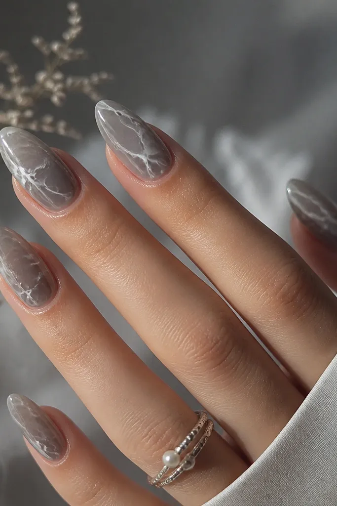 Pearl grey almond nails with a cool, smoky base. The outer half has thin shell vein lines that look like delicate cracks, highlighted with white-pearl on the top edges.