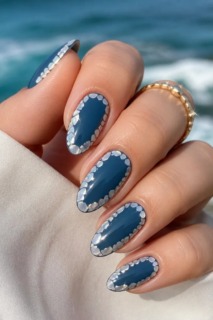 Ocean blue almond nails with a deep, glossy base. The outer edge has a mosaic-like shell outline made from small curved tiles in pearl white, with a few shimmer highlights.